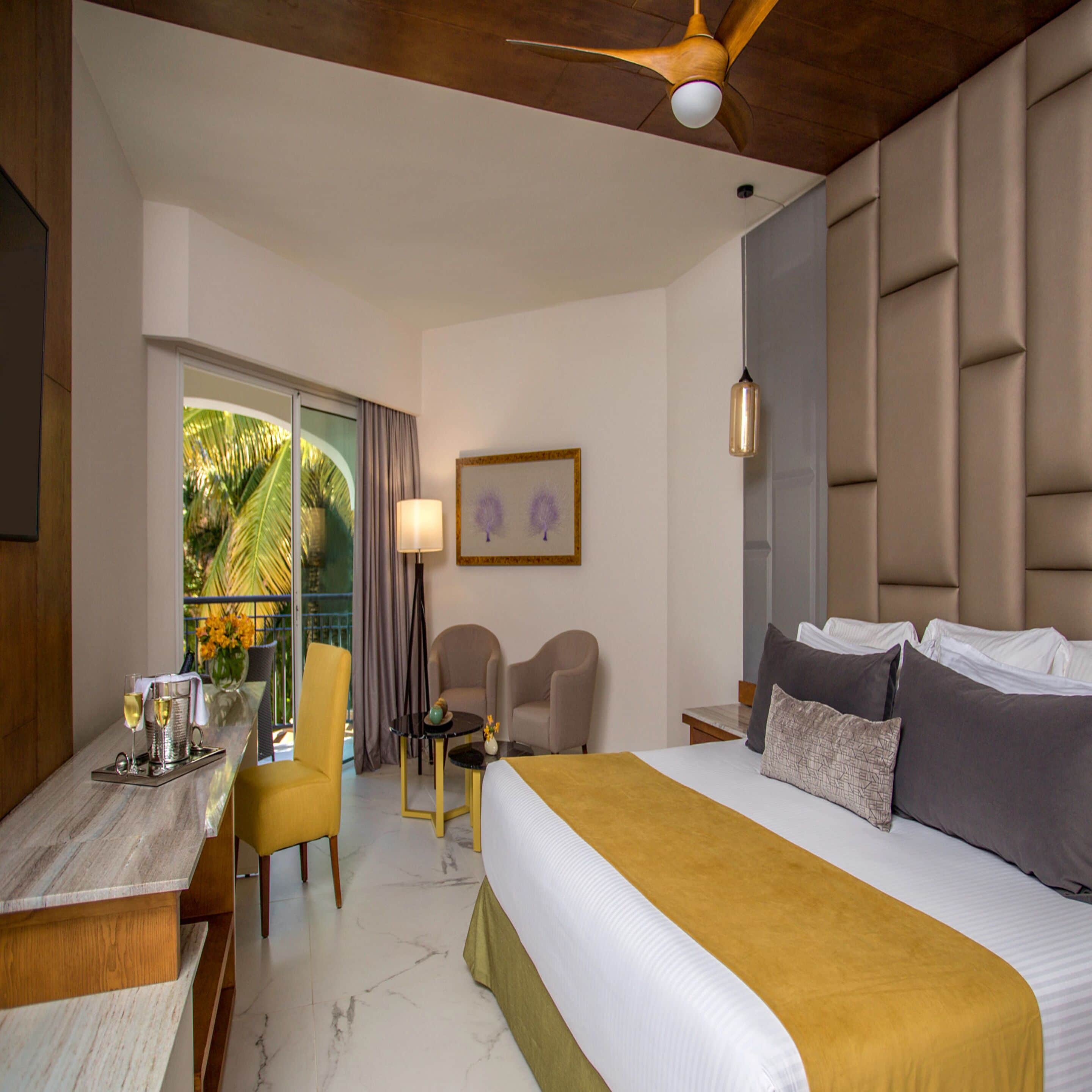 A hotel room with a bed, a desk, two chairs, a ceiling fan, and a window with a view of palm trees.