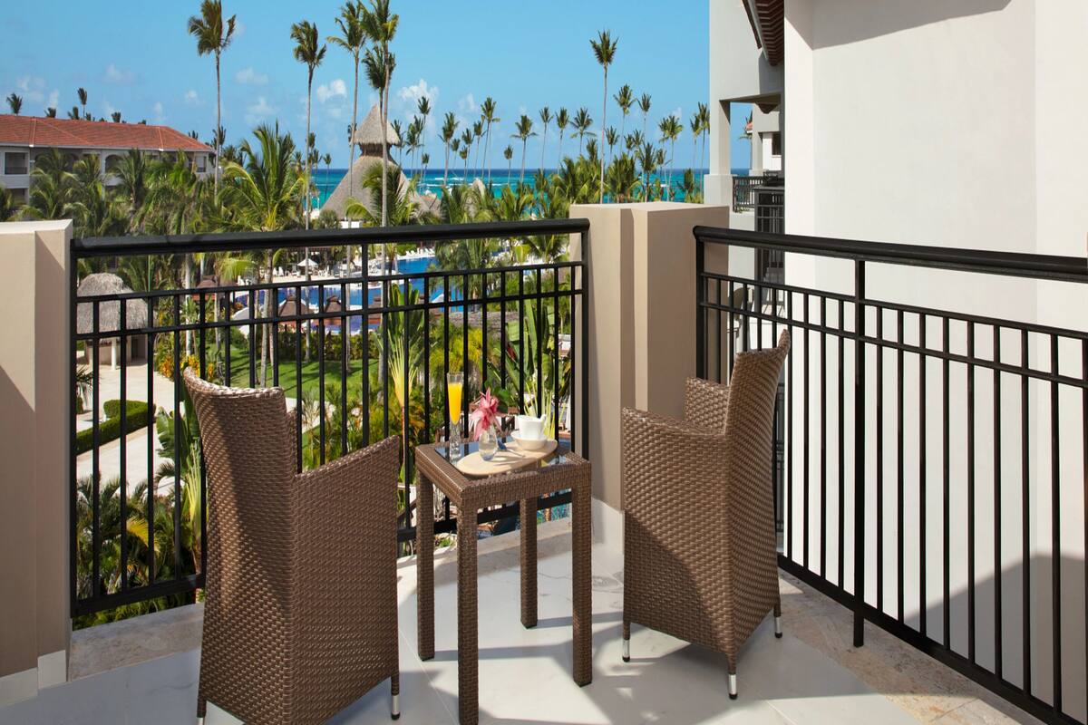 A balcony with wicker furniture, a view of a pool and palm trees, and a building with a red roof.