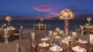 A beachside dining setup with round tables, gold chairs, and floral centerpieces against a twilight sky.