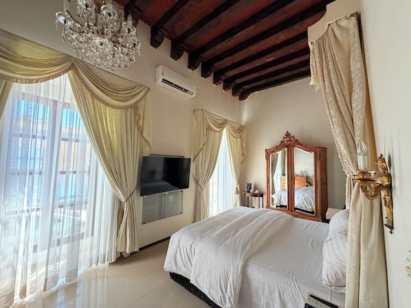 A bedroom with a large bed, a TV mounted on the wall, a mirror, and a chandelier.