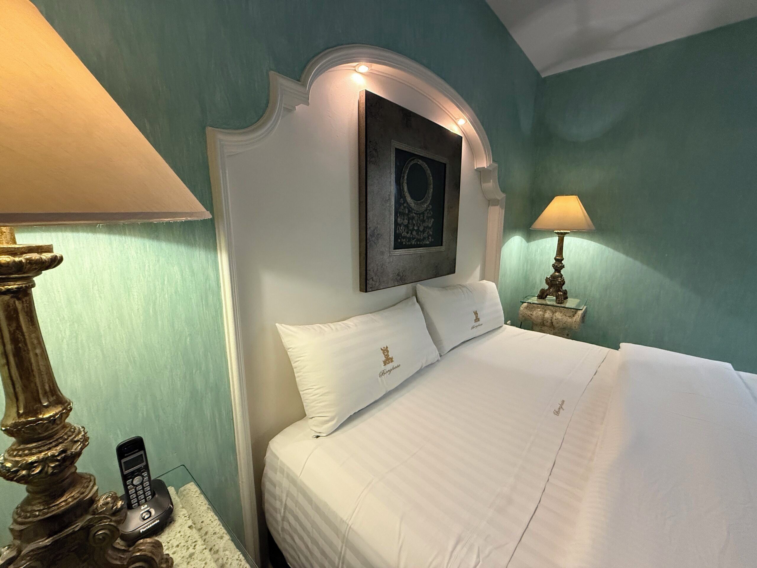 Quarto de hotel com uma cama, duas luminárias de cabeceira, um objeto decorativo na parede e um telefone na mesinha de cabeceira.