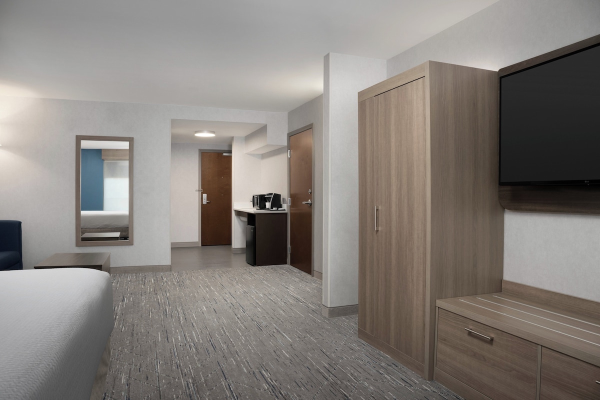 A hotel room with a large bed, a TV mounted on the wall, a wardrobe, and a small kitchenette area.