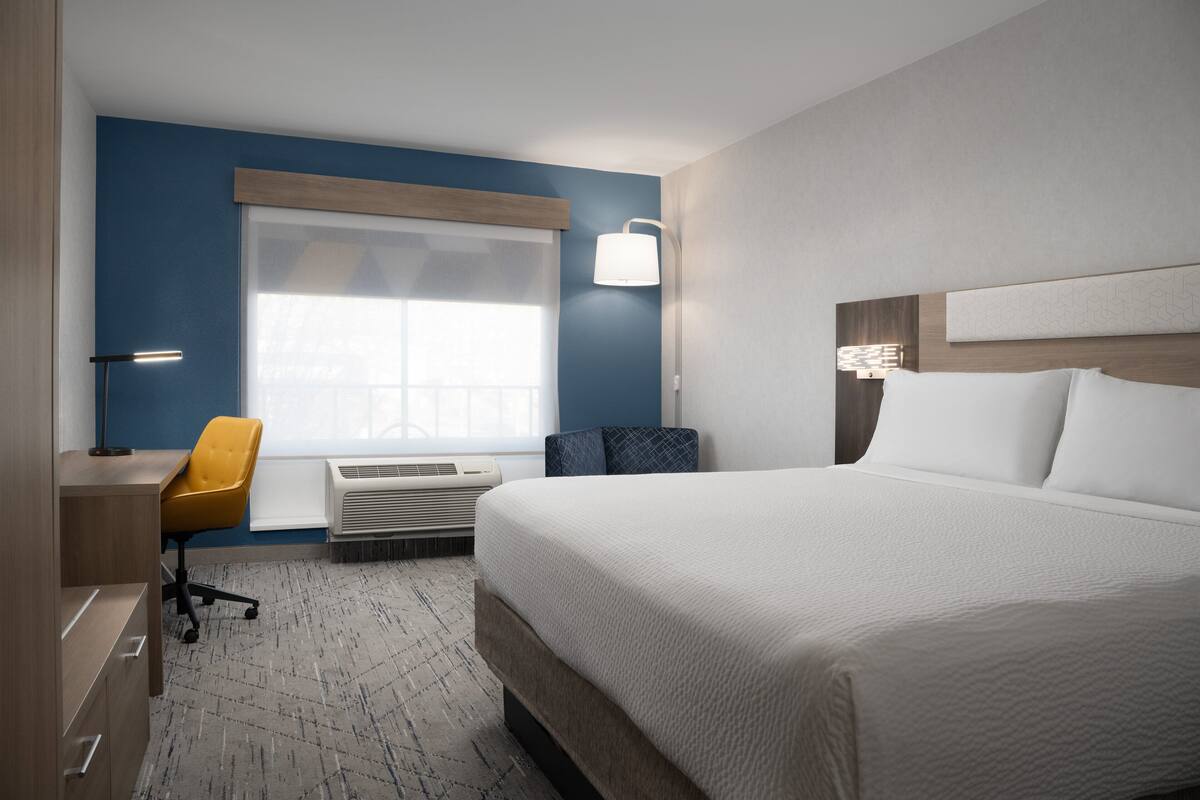 A hotel room with a large bed, a desk with a chair, a standing lamp, and a window with blinds.