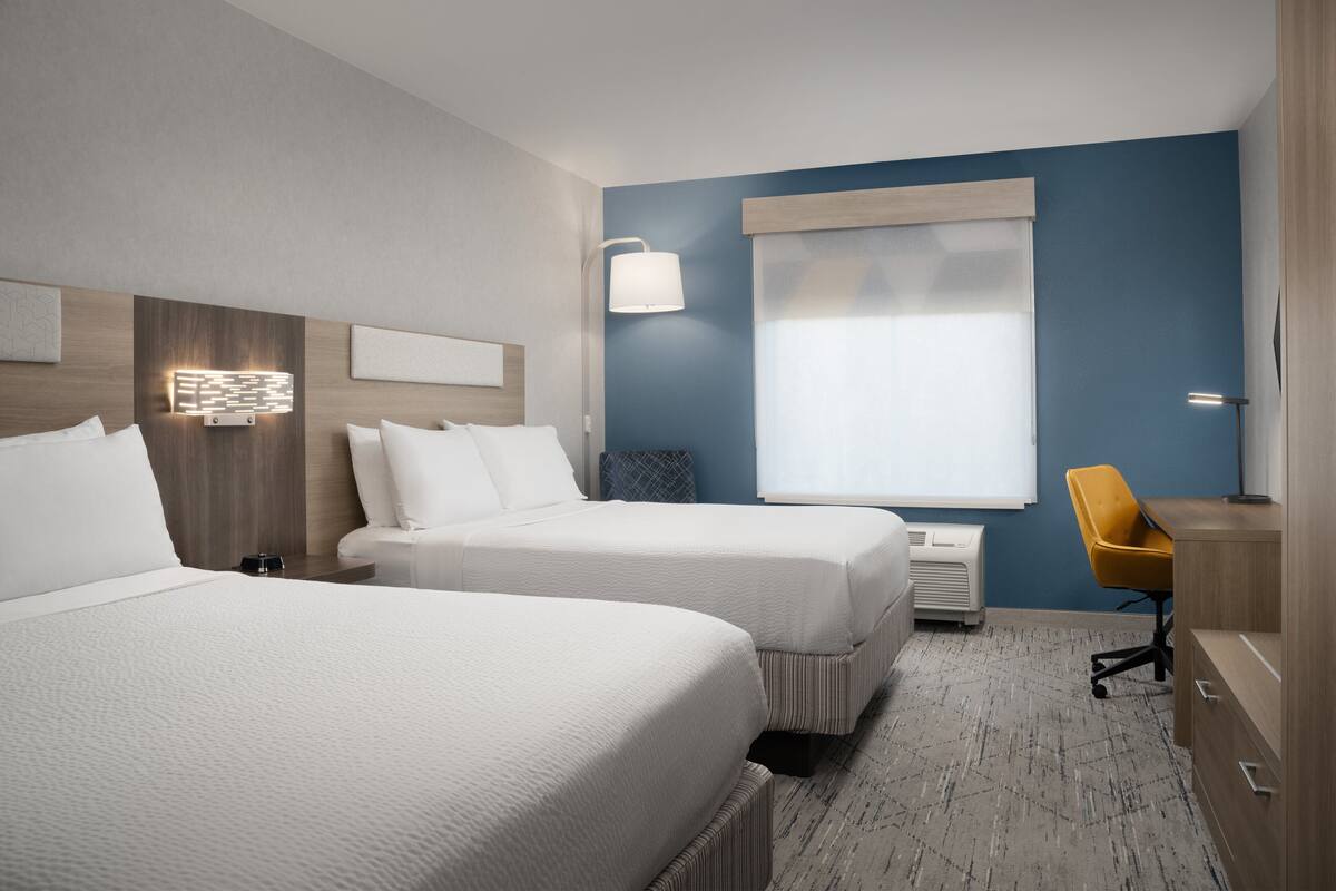 A hotel room with two beds, a desk, a chair, and a large window with blinds.