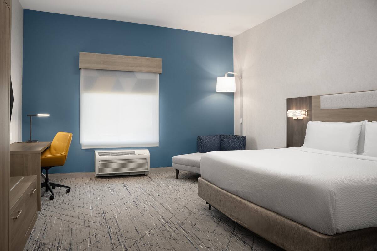 A hotel room with a large bed, a desk with a chair, a window with blinds, a wall-mounted lamp, and a wall heater.
