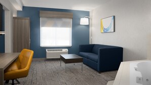 Una habitación de hotel moderna con pared azul decorativa, un sofá, una mesita, un sillón amarillo, una bañera blanca y una ventana con persianas.
