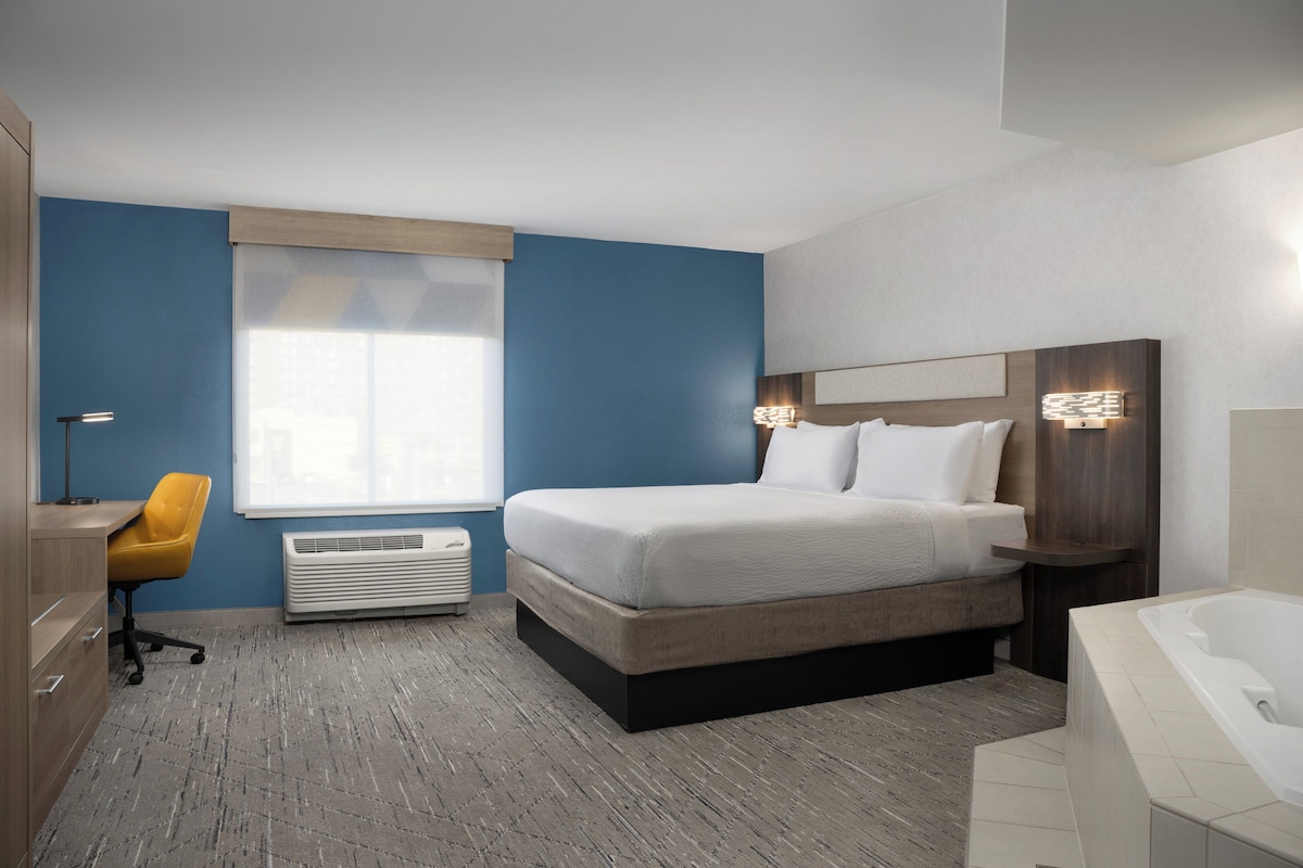 A modern hotel room with a large bed, a desk, a chair, a window, and a bathroom with a bathtub.