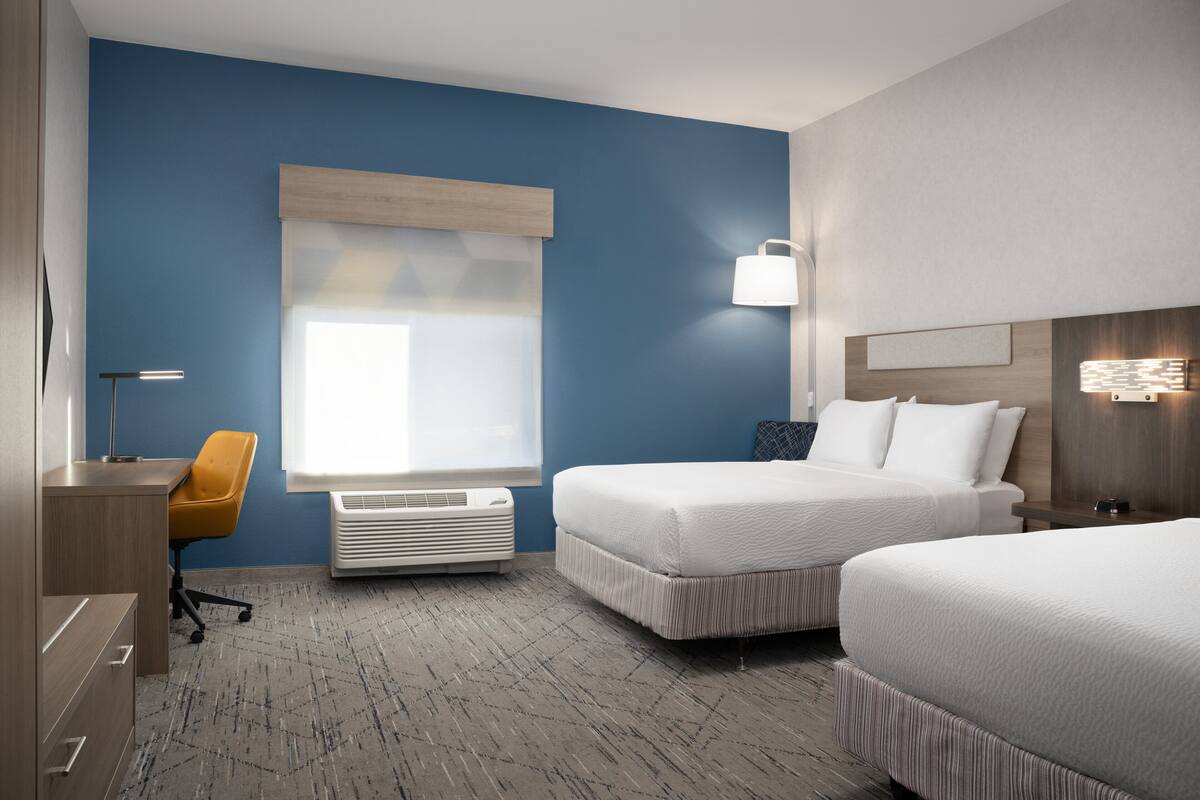 A hotel room with two beds, a desk, a chair, a window with blinds, and a wall-mounted lamp.