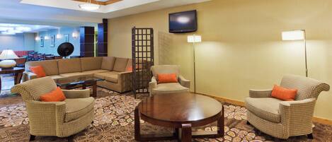 A hotel lobby with a sectional sofa, armchairs, a coffee table, a patterned carpet, a television, and a floor lamp.