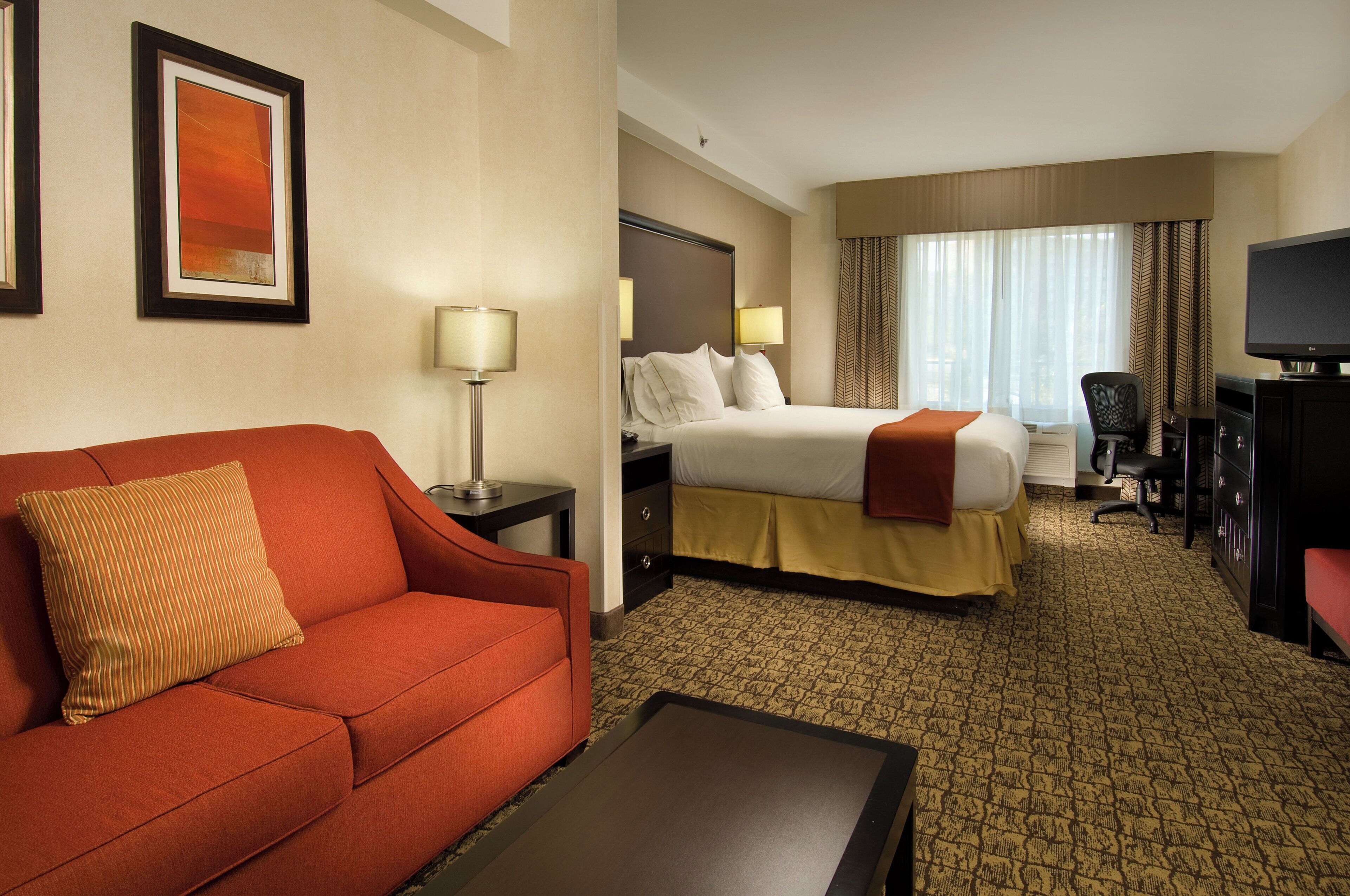 A hotel room with a red sofa, a bed with white bedding, a desk with a lamp, a television, and a patterned carpet.