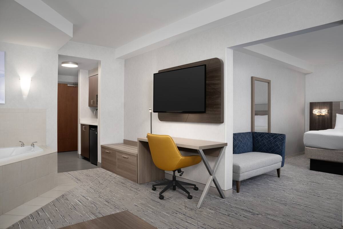 A modern hotel room with a large bed, a desk with a chair, a flat-screen TV, a bathroom with a sink and toilet, and a mirror.