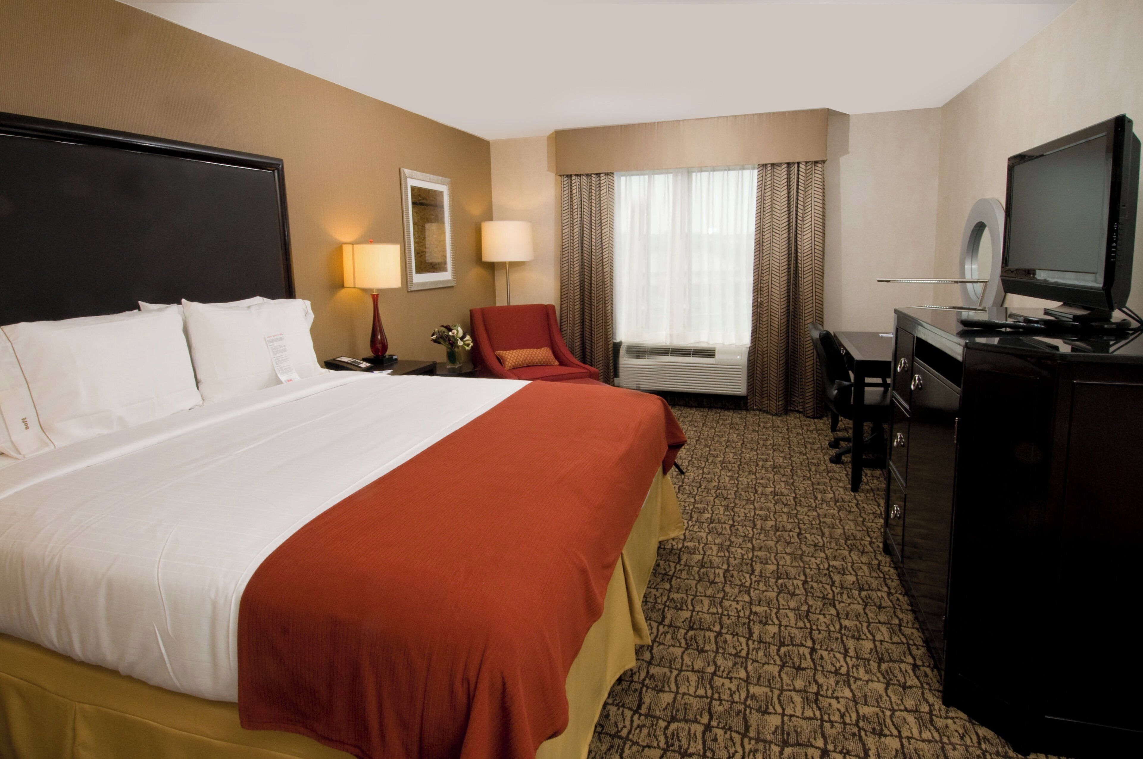 Quarto de hotel com uma cama grande, uma escrivaninha, televisão e janela com cortinas.