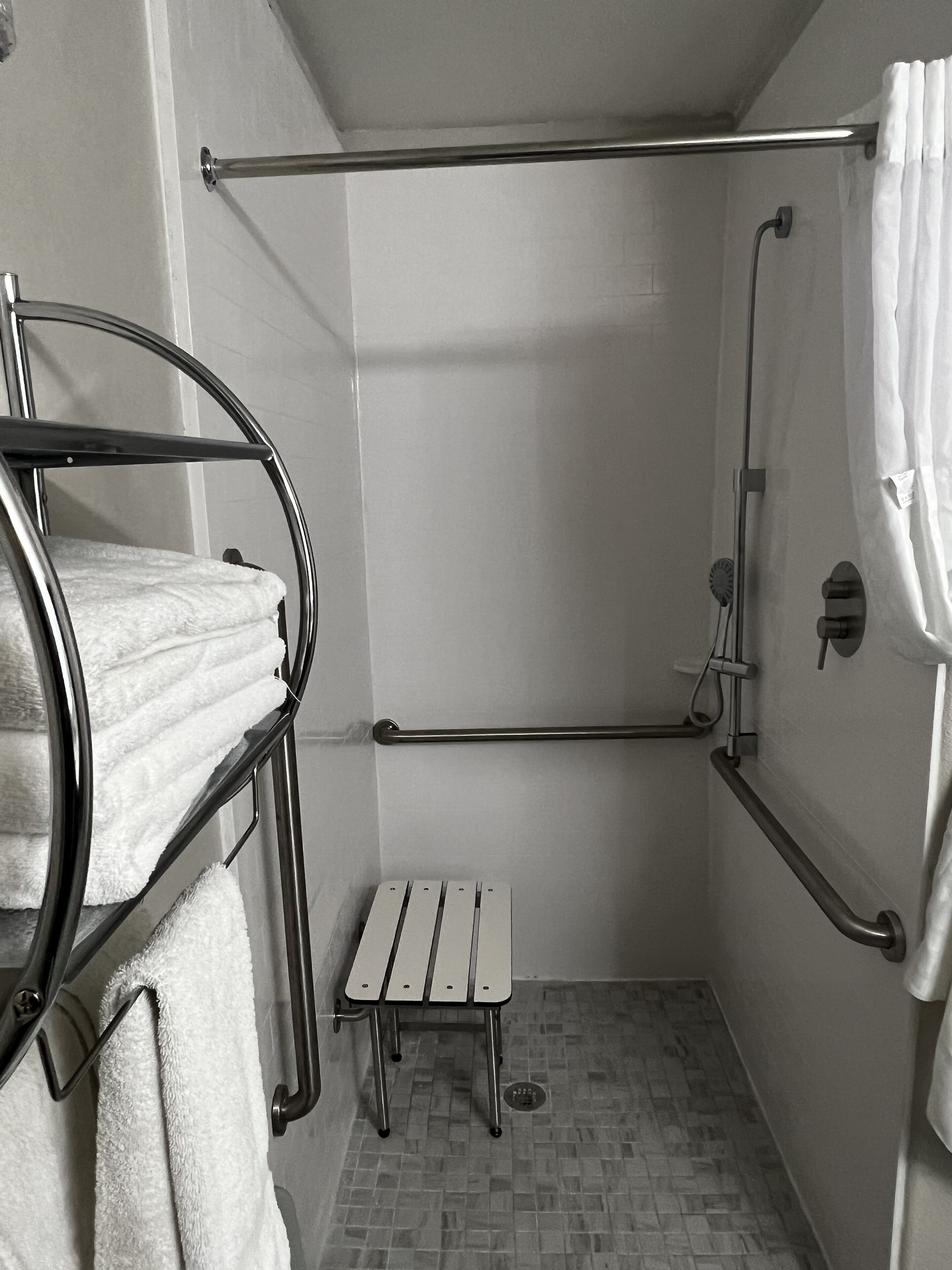 A small, enclosed shower area with a metal grab bar, a step stool, and a towel rack with towels.