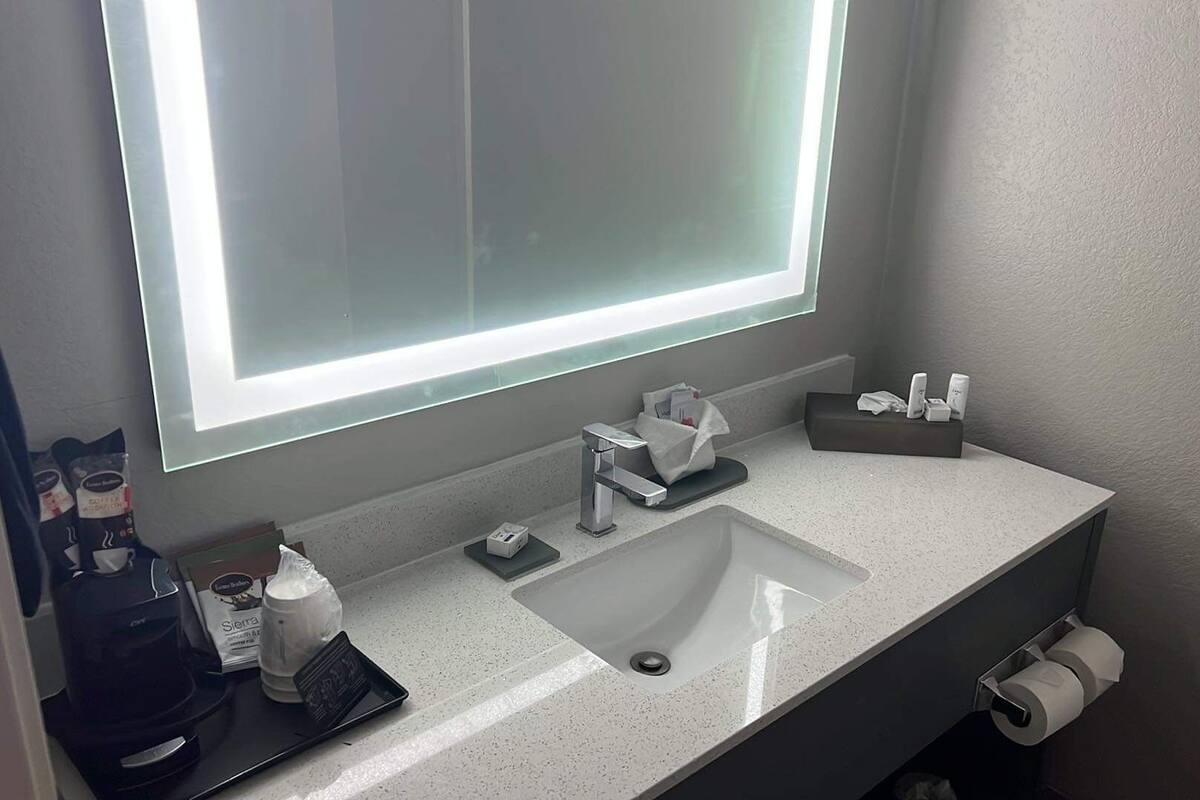 A modern bathroom with a large mirror, a sink, and a toilet. There are toiletries on the counter and a trash bin.