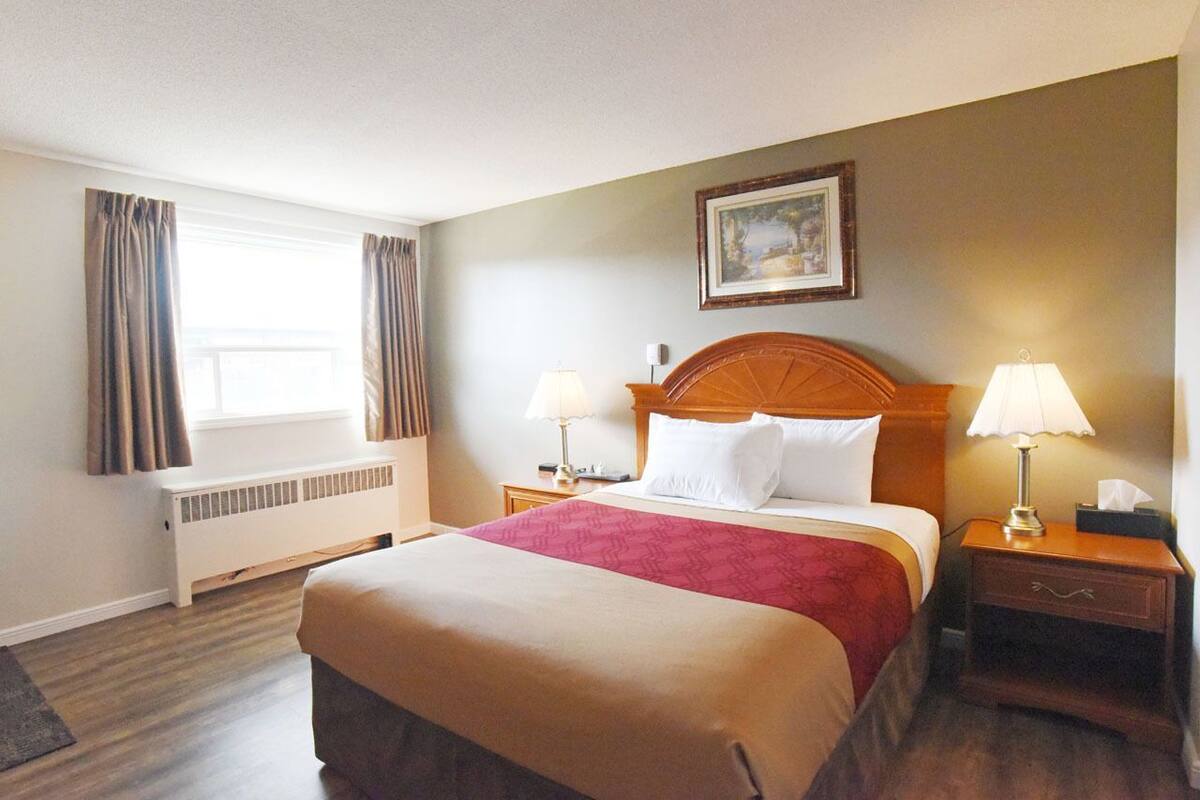 Una habitación de hotel con cama, lámparas de noche, radiador, ventana con cortinas y un cuadro en la pared.