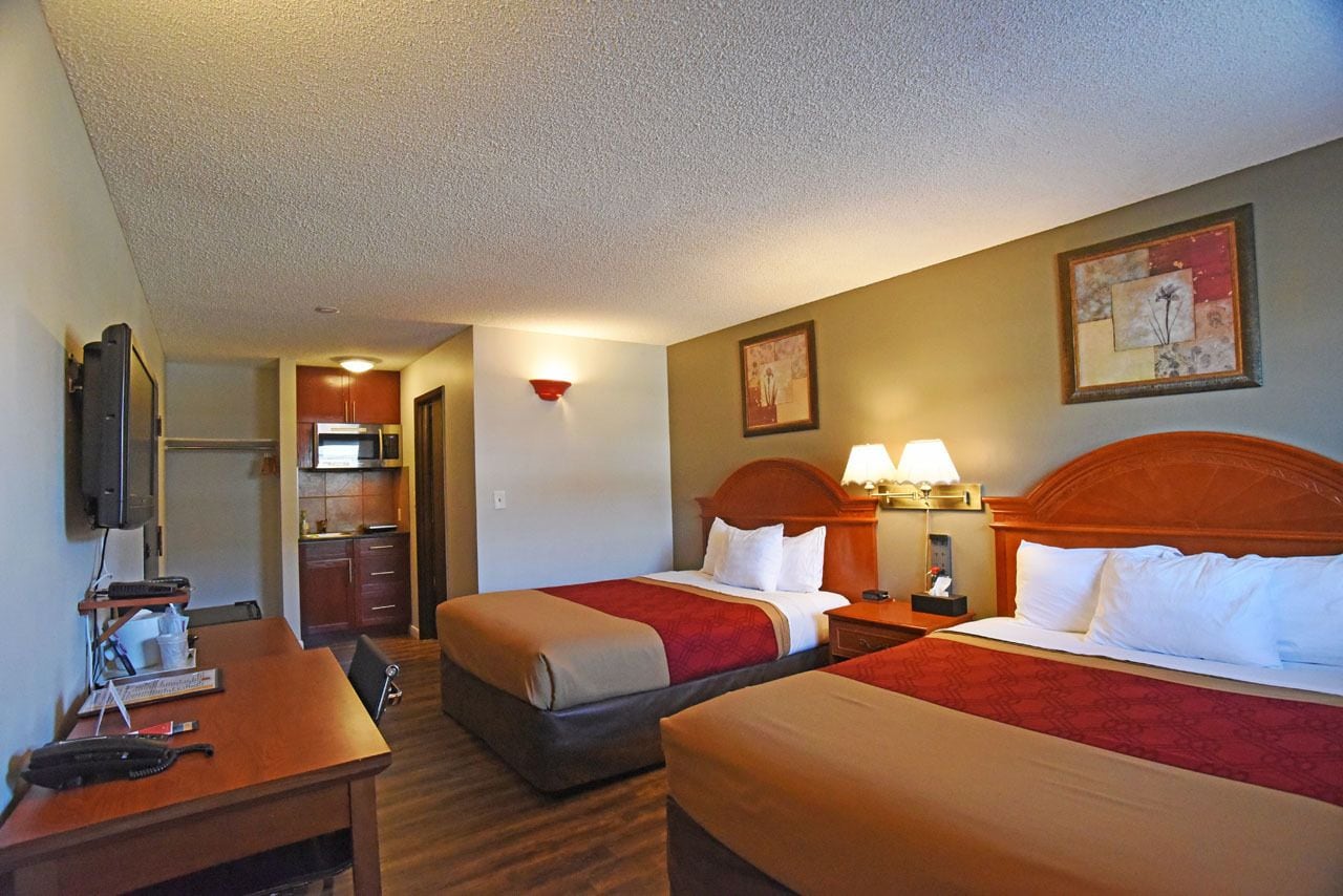 A hotel room with two beds, a desk, a television, a kitchenette, and artwork on the walls.