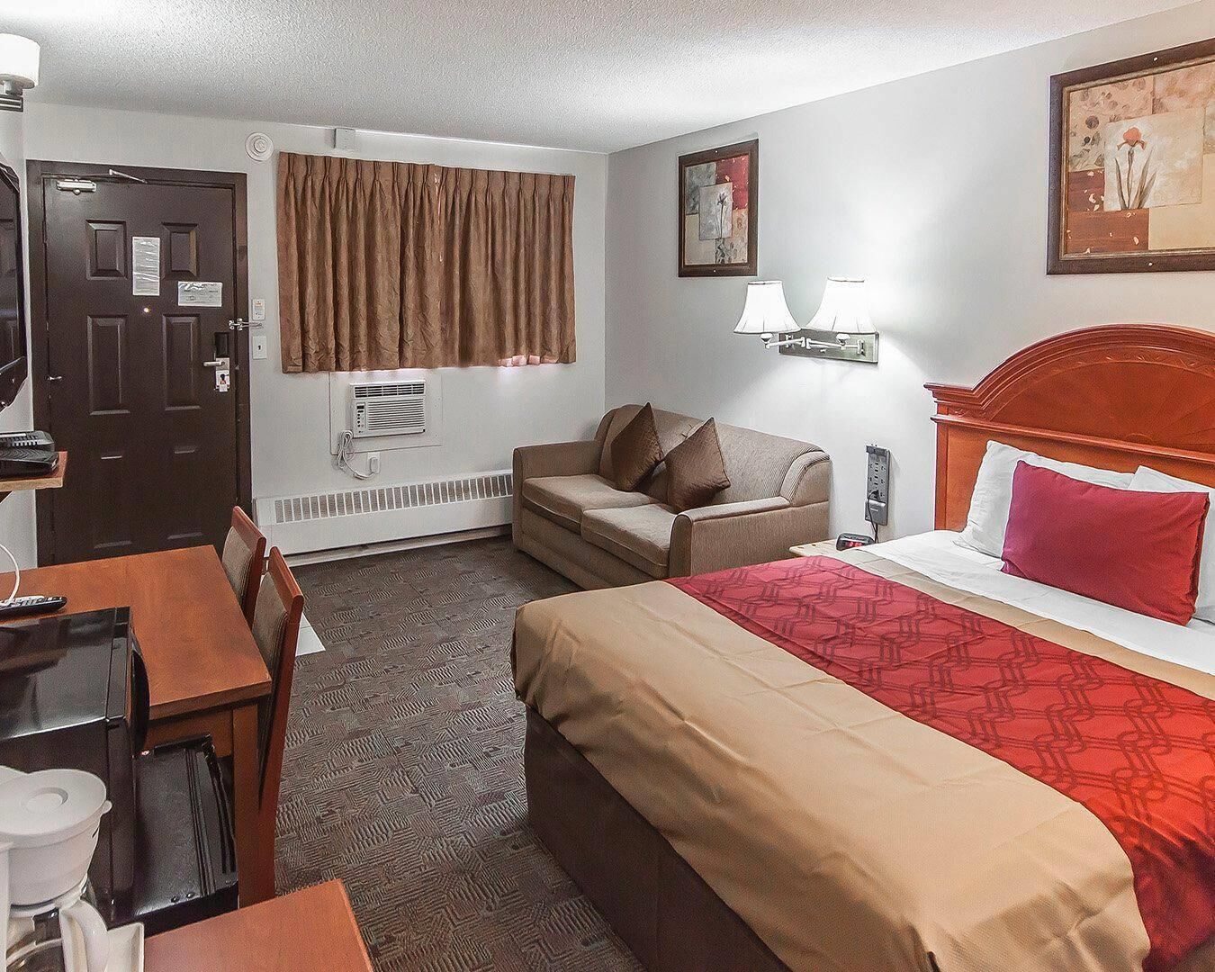 A hotel room with a bed, a desk, a chair, a couch, a window with curtains, a wall-mounted lamp, and a painting.