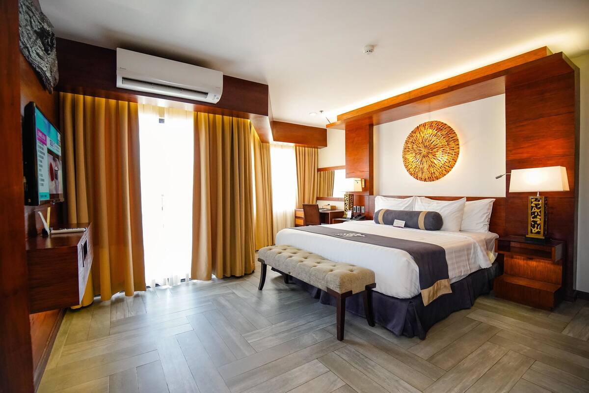 A hotel room with a large bed, a bench, a desk, and an air conditioning unit.