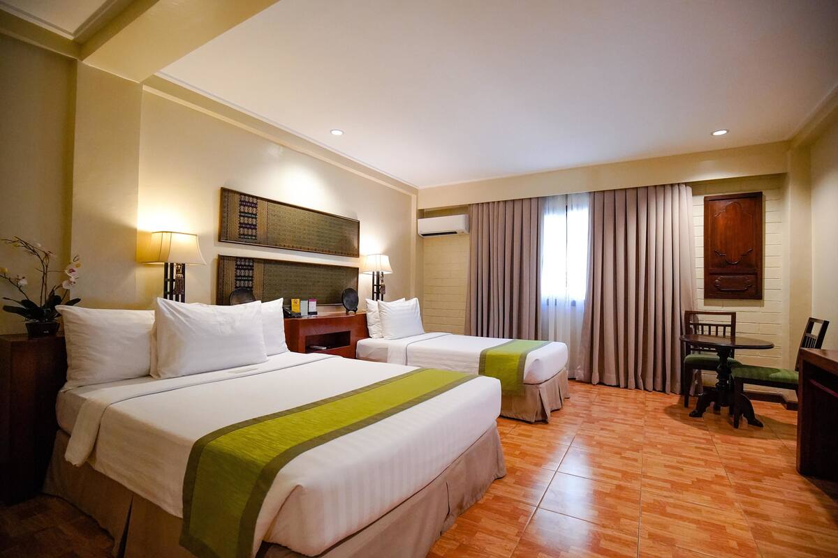 A hotel room with two beds, a desk, a chair, a lamp, and a potted plant.