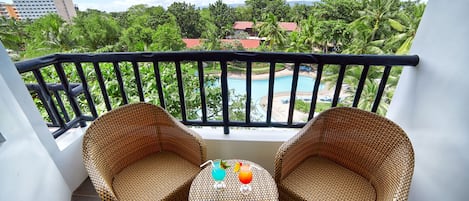 Ein Balkon mit Korbstühlen und einem kleinen Tisch, der einen Blick auf einen Pool und üppiges Grün bietet.
