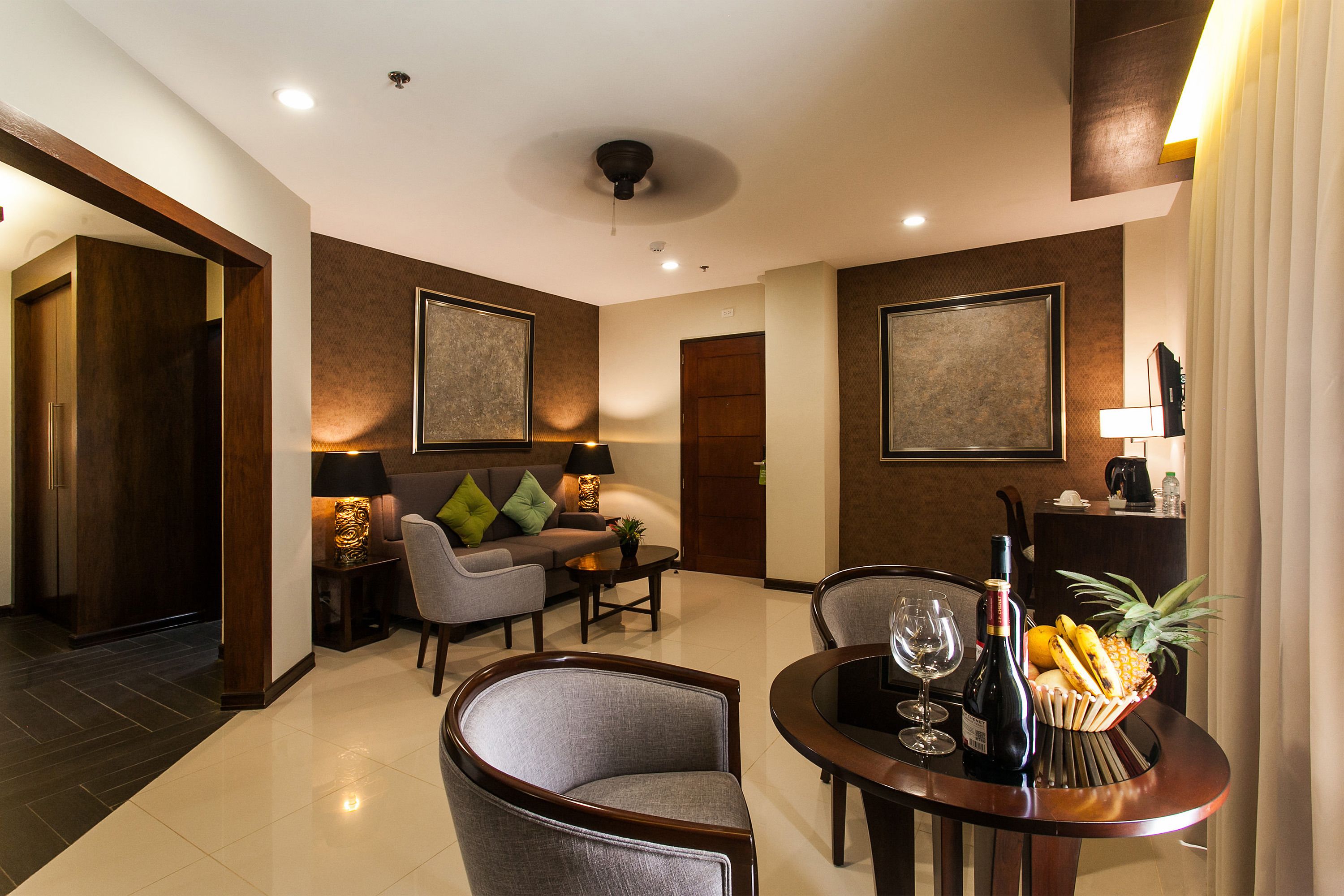 Ein modernes Hotelzimmer mit einer Couch, zwei Sesseln, einem Esstisch mit Obstschale, Weingläsern und einer Weinflasche.