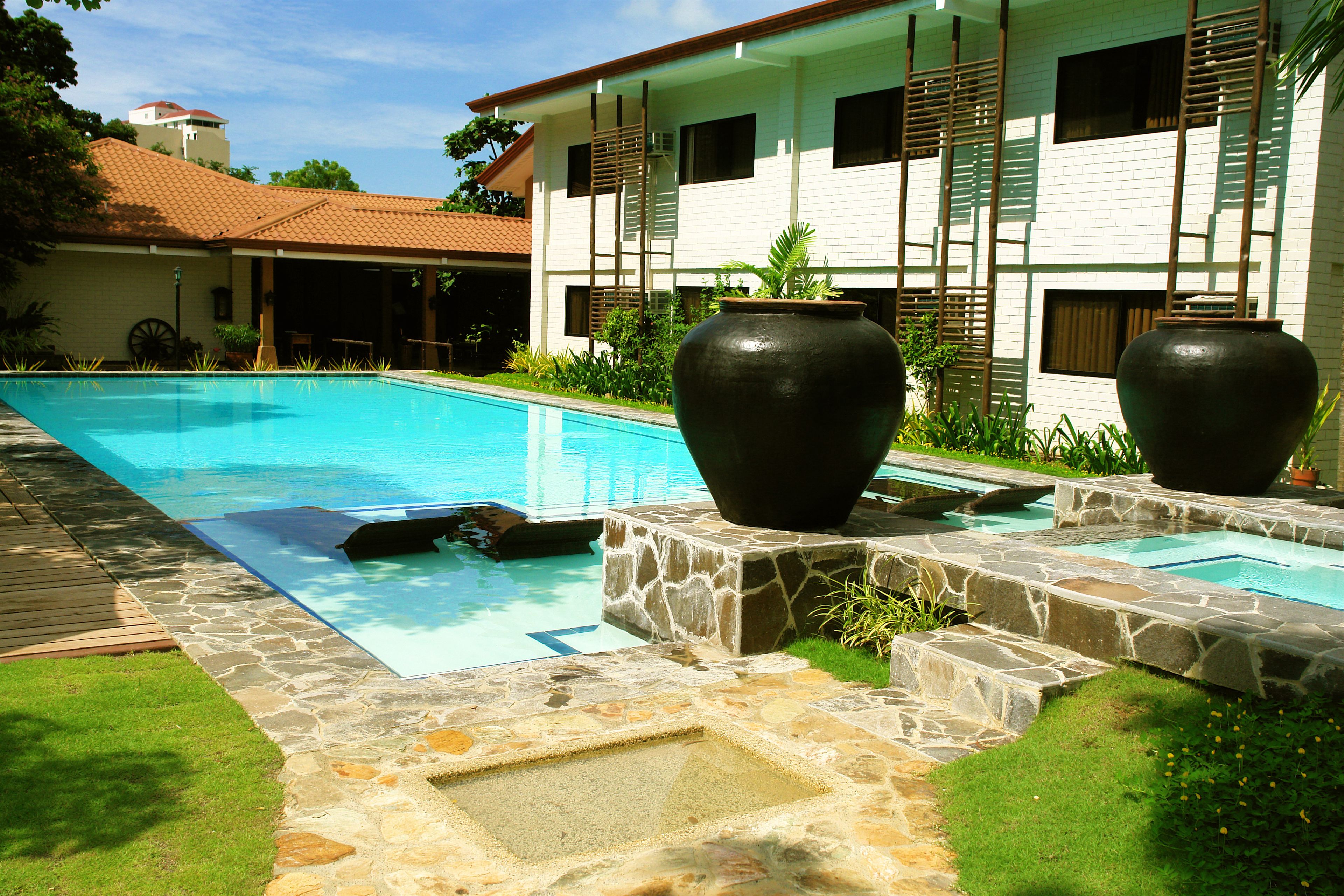 A swimming pool with stone and grass borders, large black vases, and a white building with shutters in the background.