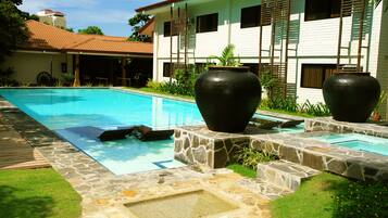 A swimming pool with stone and grass borders, large black vases, and a white building with shutters in the background.