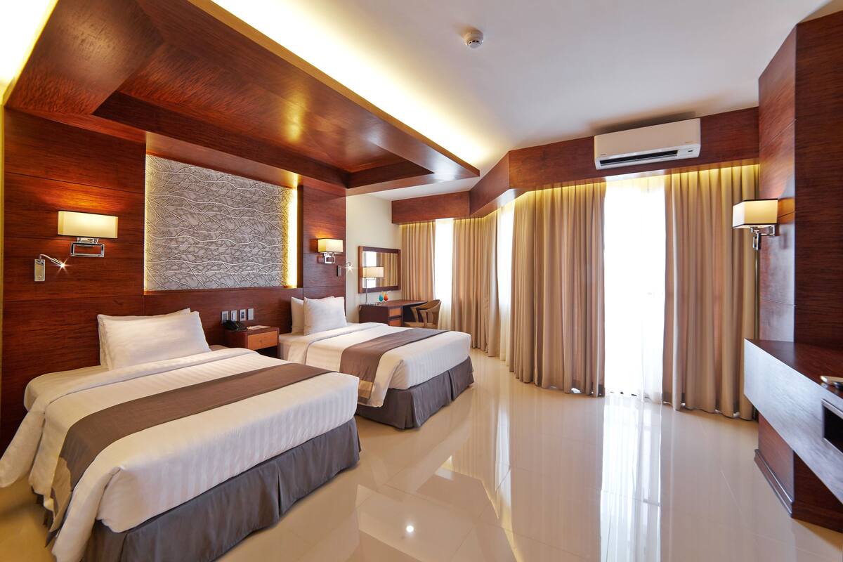 A hotel room with two beds, a desk, a chair, a TV, and a large window with curtains.