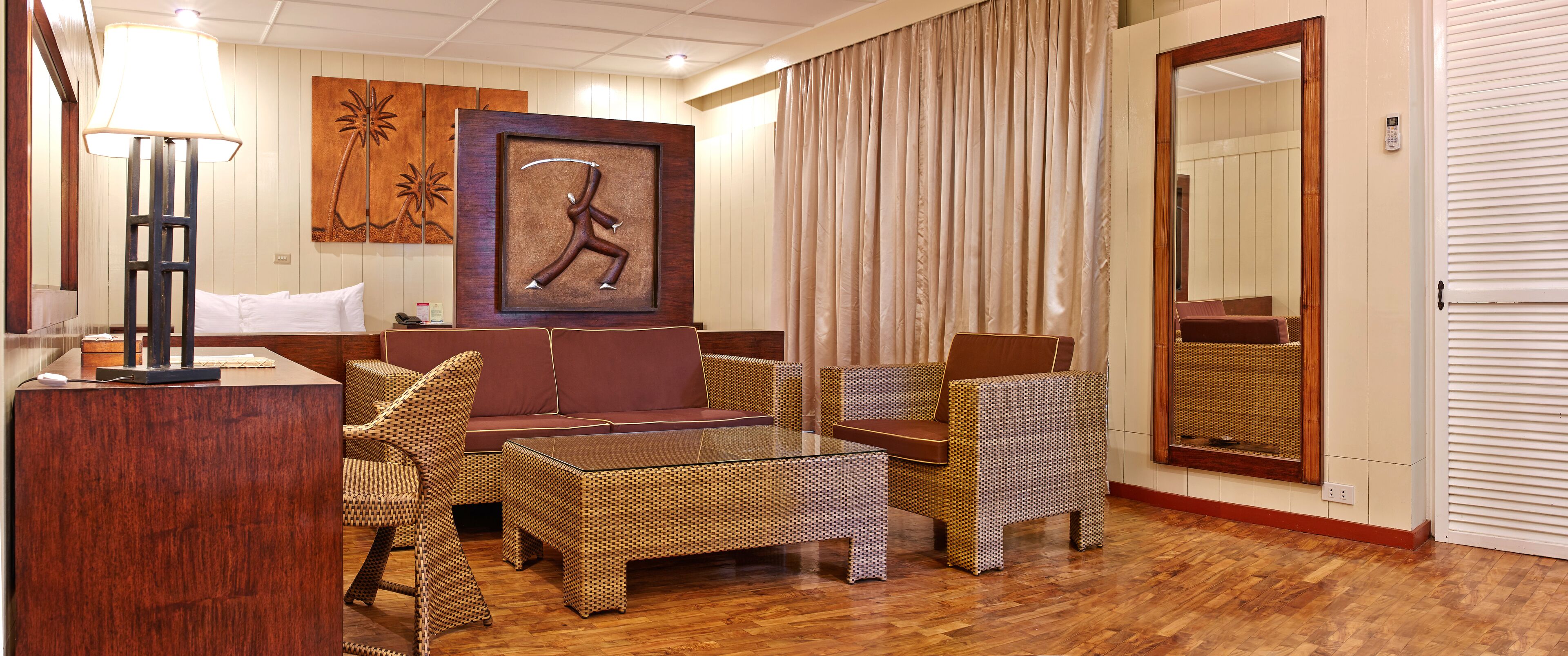 Una habitación de hotel con piso de madera, un sofá, dos sillas de mimbre, una mesa de centro de vidrio, una lámpara, un cuadro y un espejo grande.