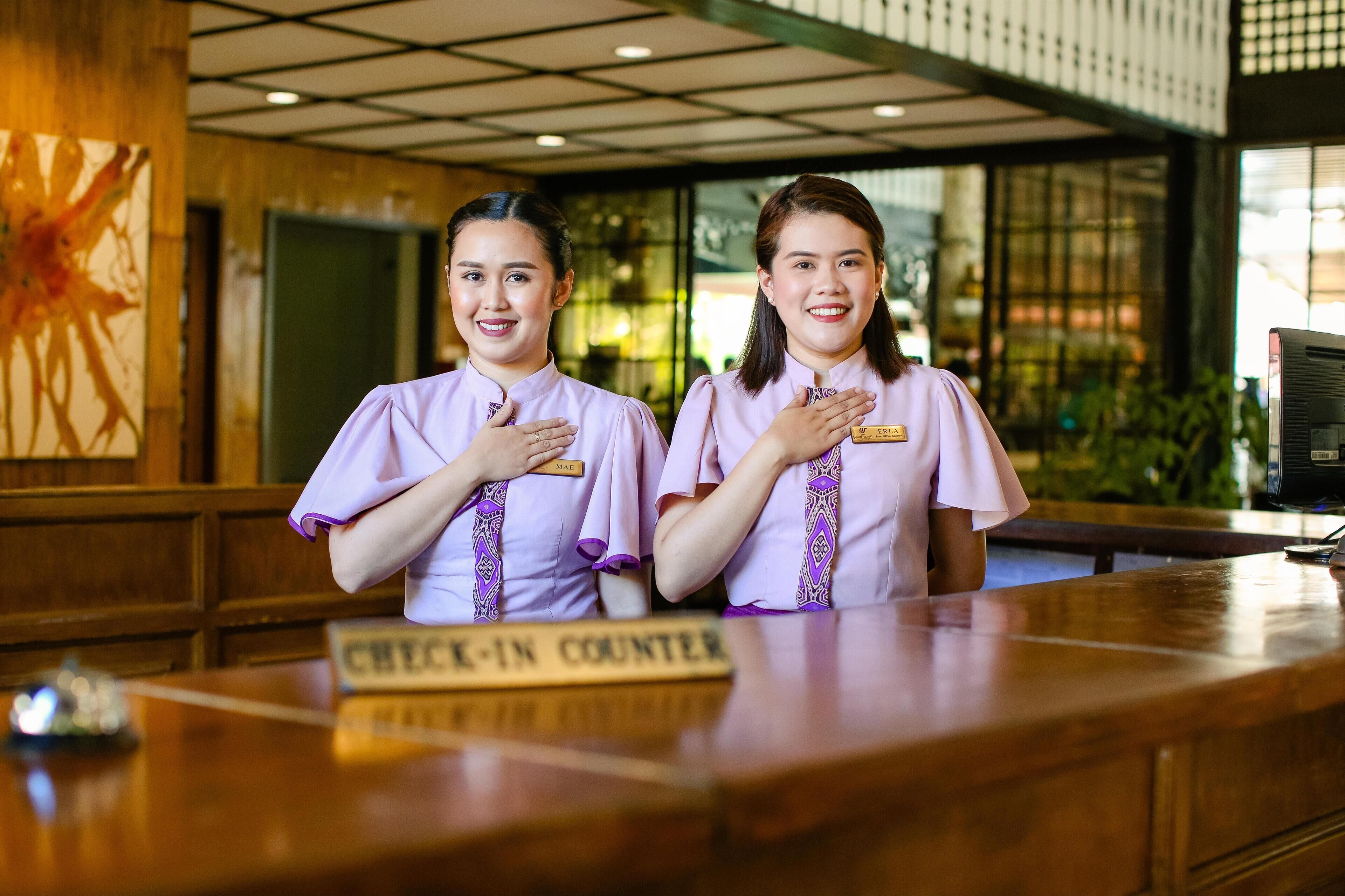 Two receptionists at a check-in counter with a name tag and a sign that reads "CHECK-IN COUNTER".