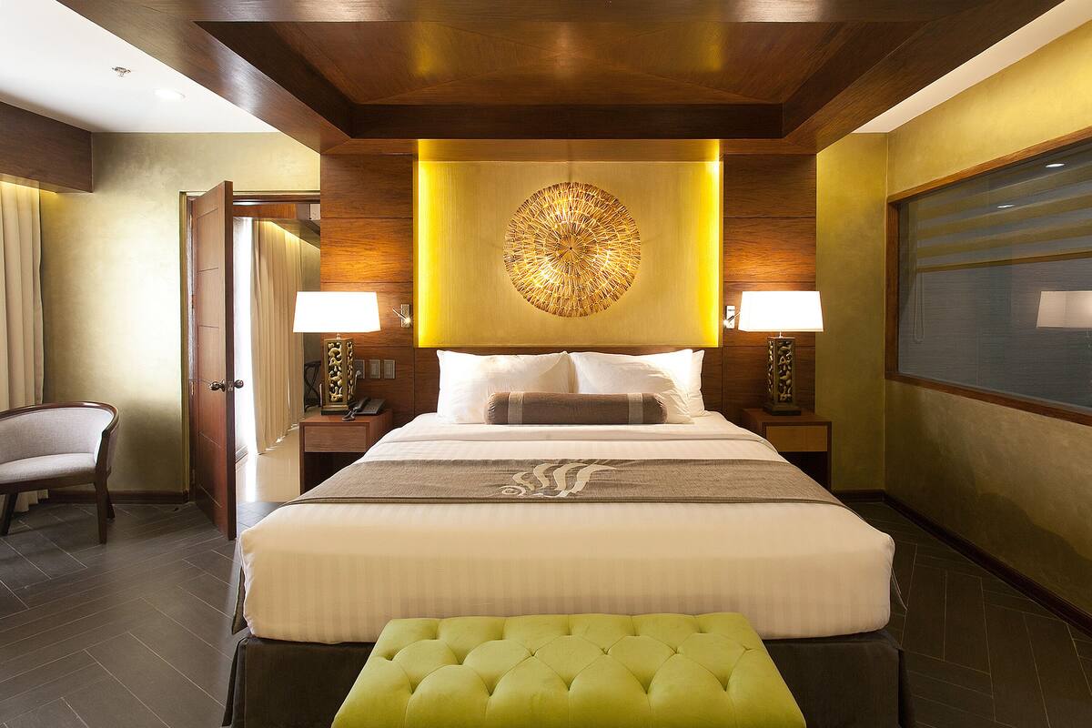 A hotel room with a large bed, a green ottoman, a chair, and a wall-mounted lamp.