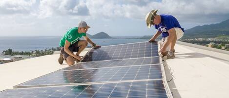 Two individuals installing solar panels on a rooftop with a view of the ocean and mountains in the background.