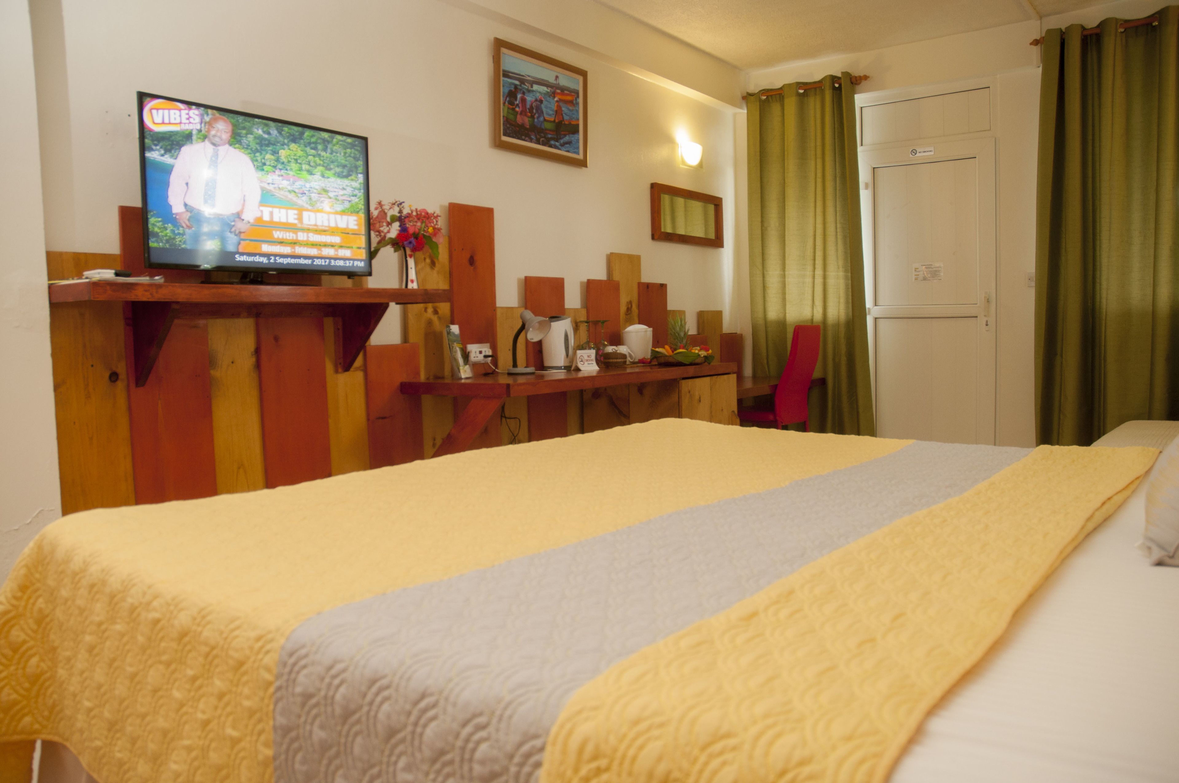 A hotel room with a bed, a television displaying a news program, a small table with a vase of flowers, and a door with curtains.