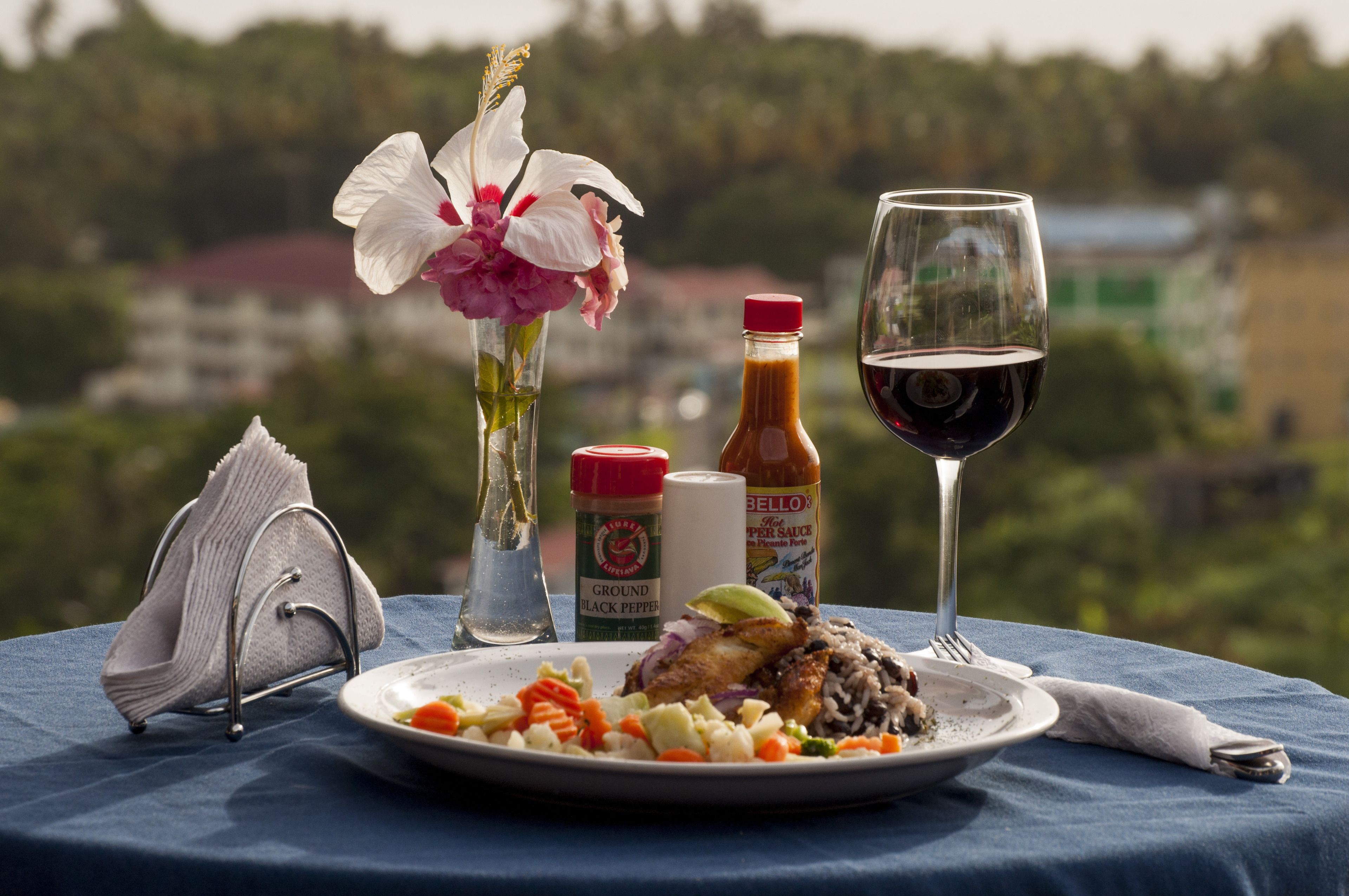 Une table avec une assiette de nourriture, un verre de vin rouge, une bouteille de sauce et un vase de fleurs.