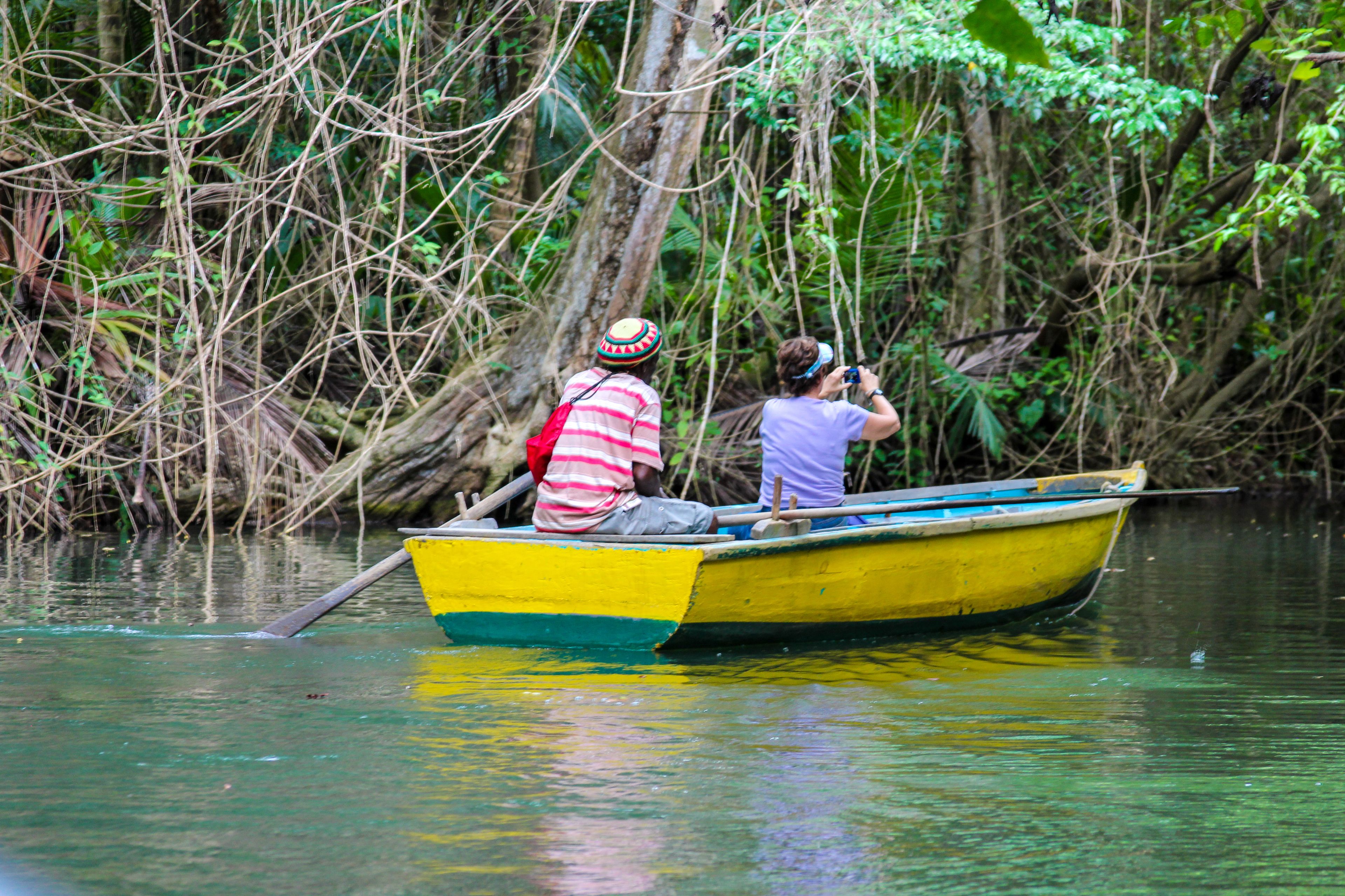 Una pequeña embarcación amarilla con dos personas, una vestida con una camisa a rayas y la otra con una camisa blanca, navegando por una vía fluvial bordeada de vegetación densa.