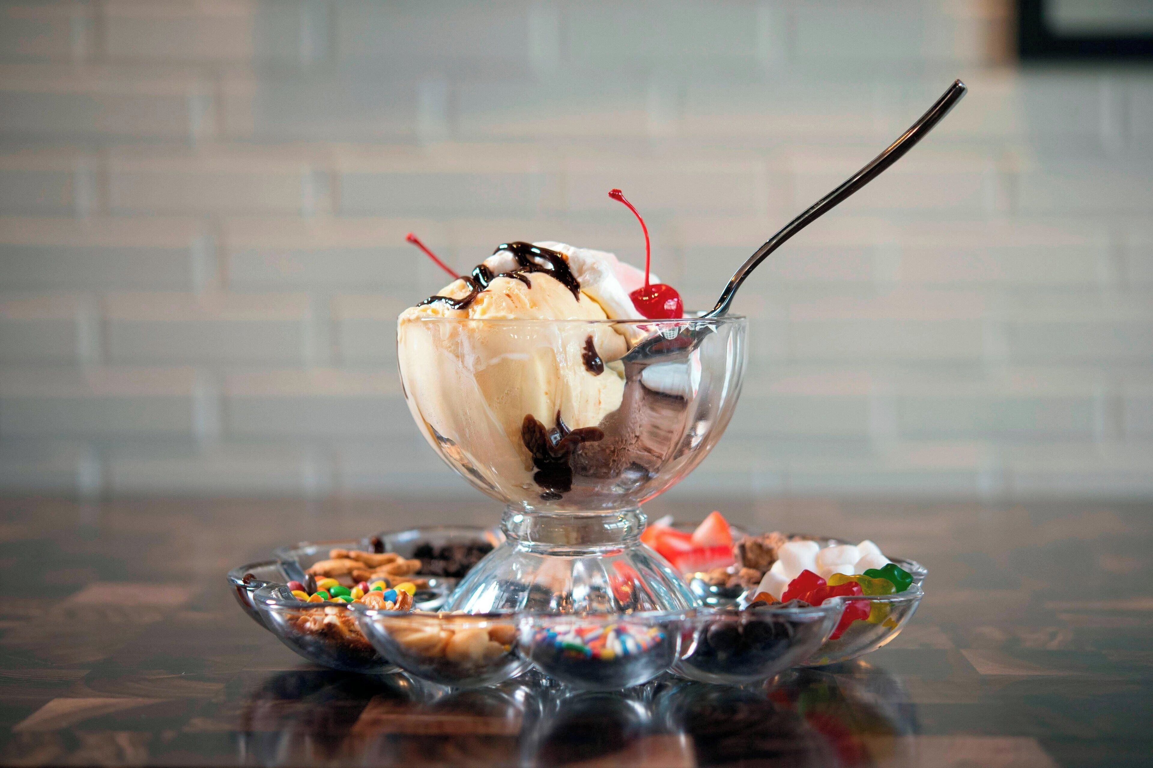 A dessert in a glass bowl with a spoon, surrounded by assorted candies and nuts on a reflective surface.