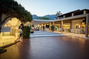 Un paseo peatonal con tiendas y mesas al aire libre, rodeado de vegetación y montañas.