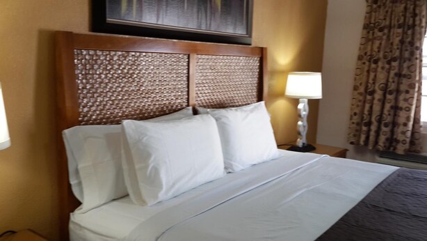 A neatly made bed with white linens and pillows, a wooden headboard with a woven pattern, a bedside lamp, and a window with curtains. - American Regency Inn (Williams)