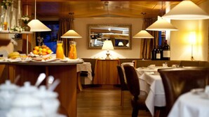 A restaurant with wooden tables and chairs, a central table with a lamp, and a view of the kitchen through a window. - Rosa Canina (Sankt Anton am Arlberg)