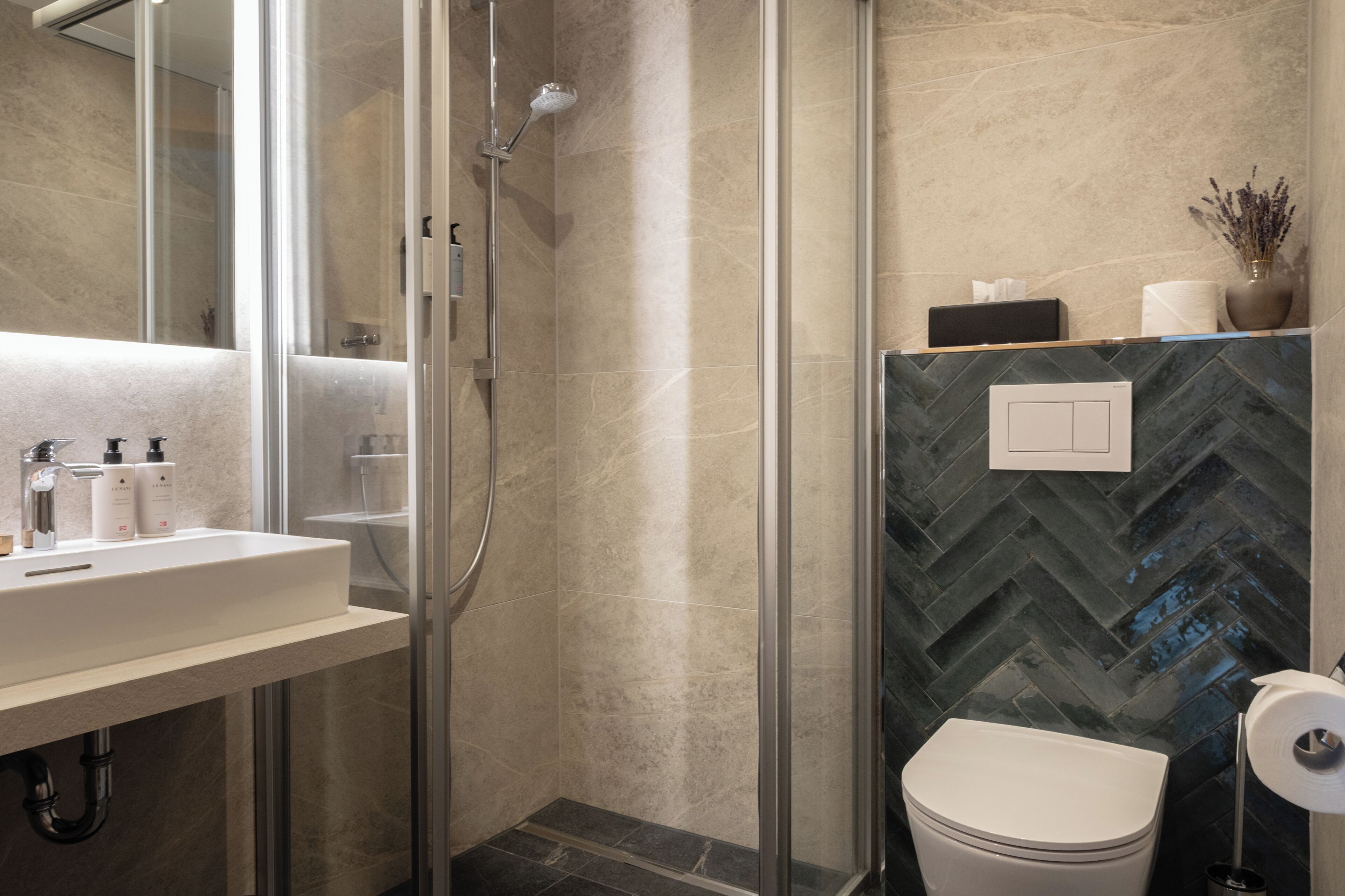 A modern bathroom with a glass-enclosed shower, a white sink with toiletries, and a herringbone-patterned tile backsplash.