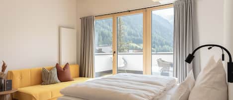 Quarto com cama, sofá, janela com cortinas e vista para montanhas.