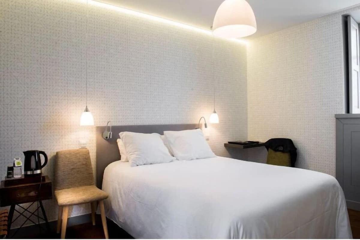 Une chambre d’hôtel avec un grand lit, deux lampes de chevet, une chaise, une petite table avec une bouilloire et une fenêtre.