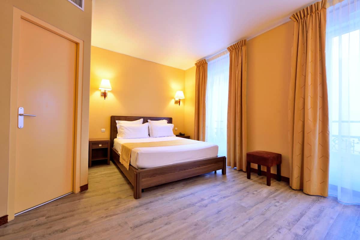 Una habitación de hotel con una cama grande, dos lámparas de noche, una mesita de madera, un taburete pequeño y una puerta.