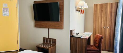 A hotel room with a bed, a desk, a chair, a TV mounted on the wall, and a wardrobe.