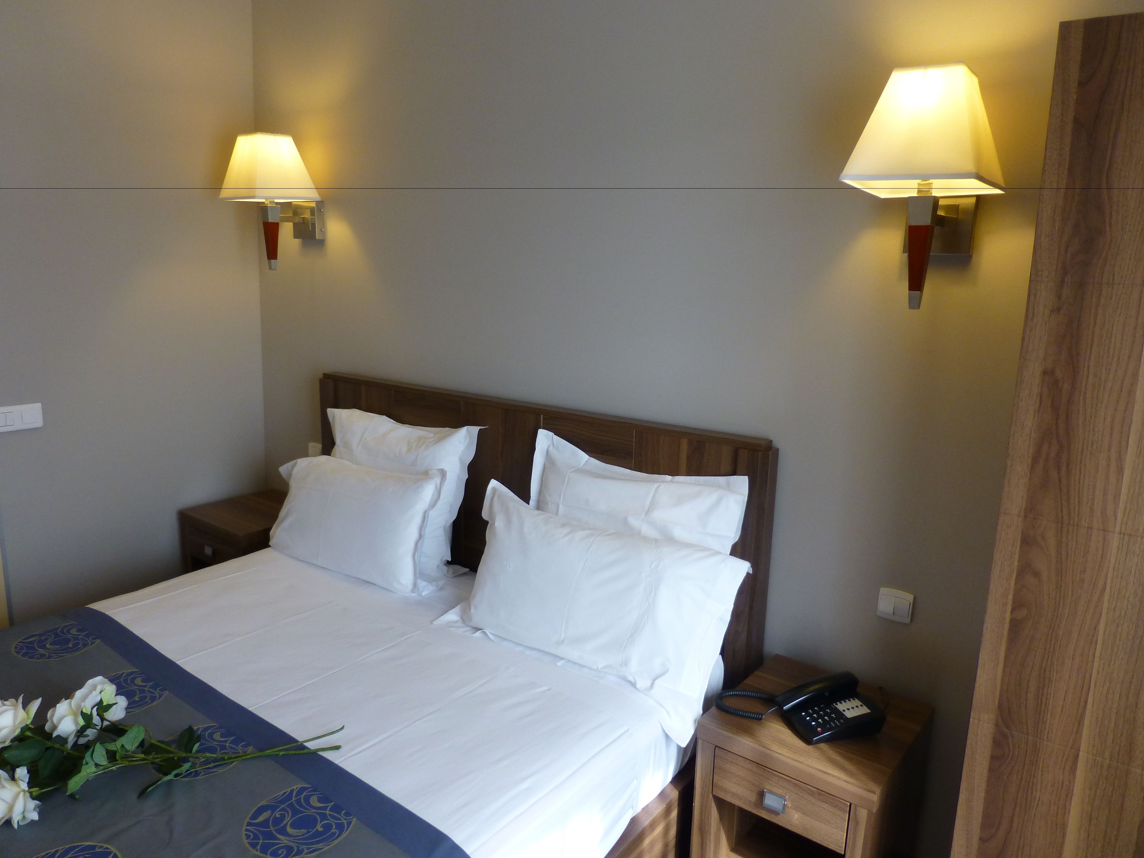 A hotel room with a neatly made bed, two bedside lamps, a wooden nightstand with a telephone, and a wall-mounted light switch.
