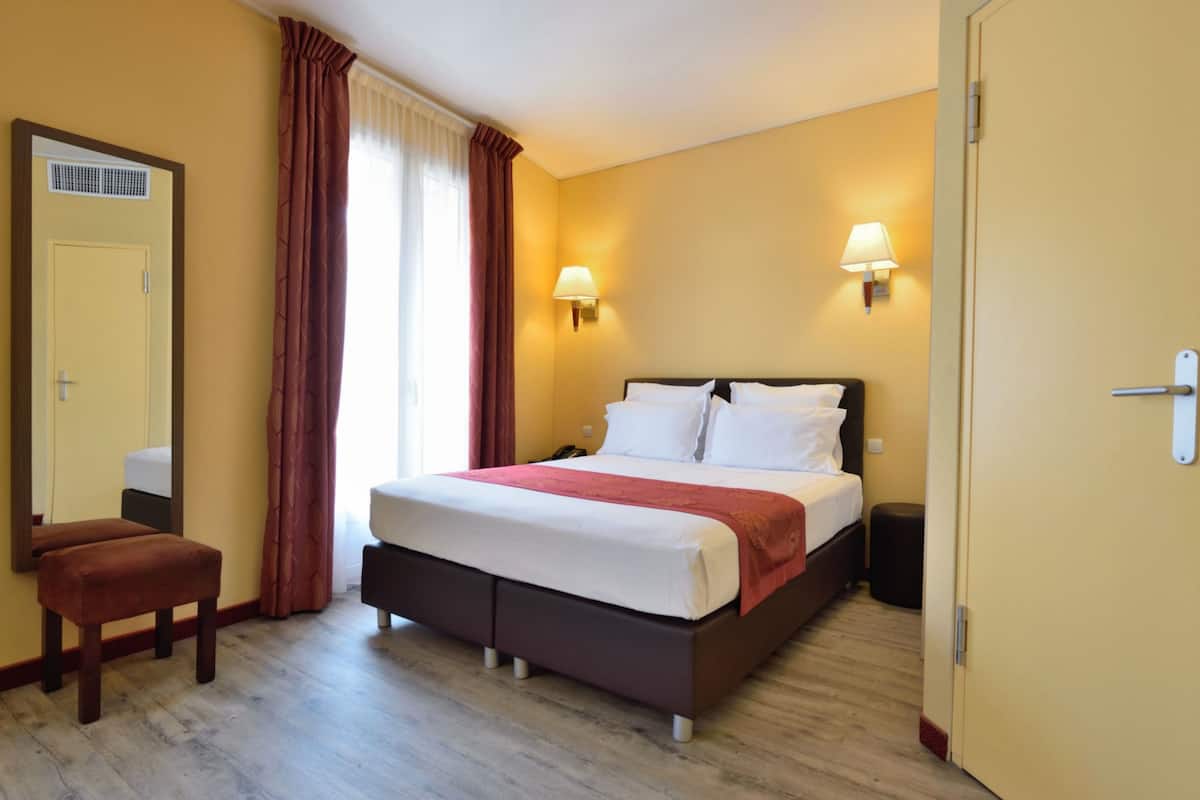 Habitación de hotel con cama, lámparas de noche, espejo, ventana con cortinas y una puerta.