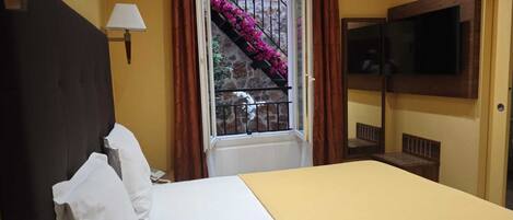 A hotel room with a bed, a TV mounted on the wall, a window with a view of stairs and flowers, and a lamp on the wall.