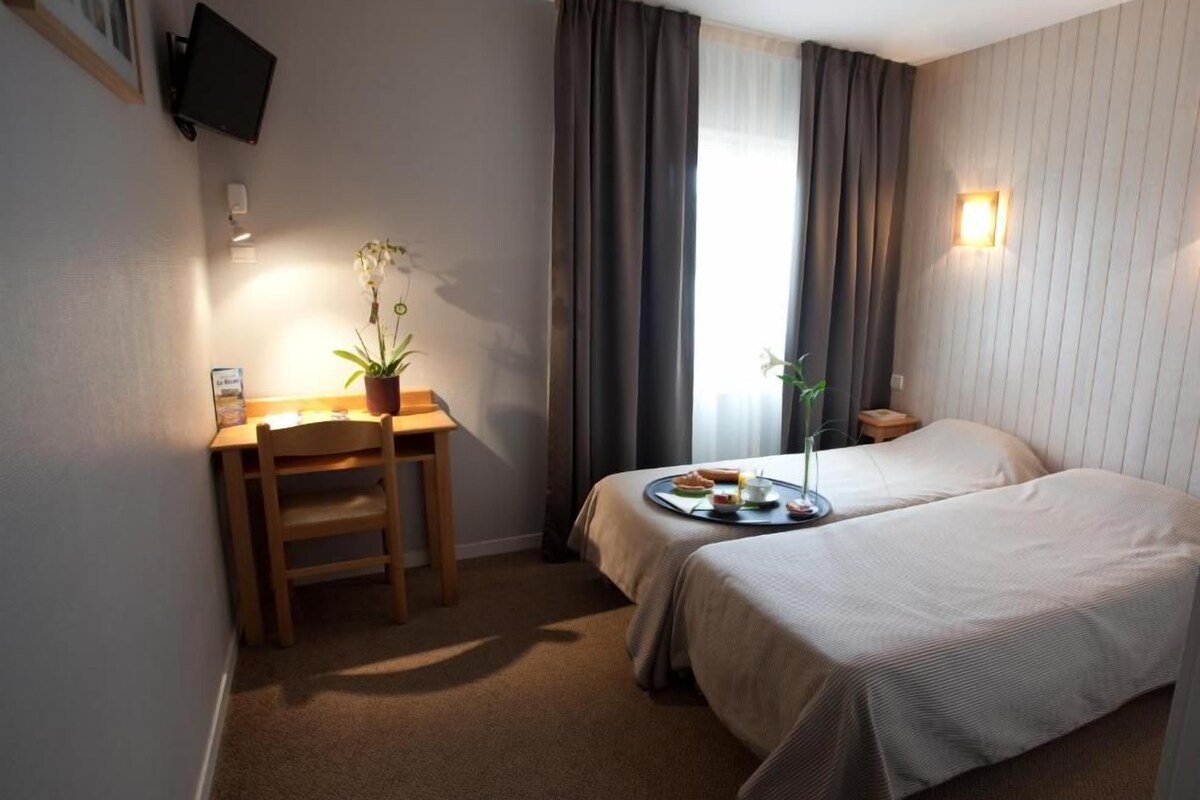 Een hotelkamer met een bureau, stoel, twee bedden, een potplant en een raam met gordijnen.