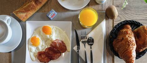 Una mesa de desayuno con huevos, tocino, cruasanes, una rebanada de pan, un vaso de jugo de naranja, una botella de leche, un salero, un molinillo de pimienta y una planta.