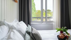 Una cama bien hecha con sábanas blancas, una ventana con cortinas, y una mesita con una cesta que contiene un croissant, una manzana y un florero con flores.